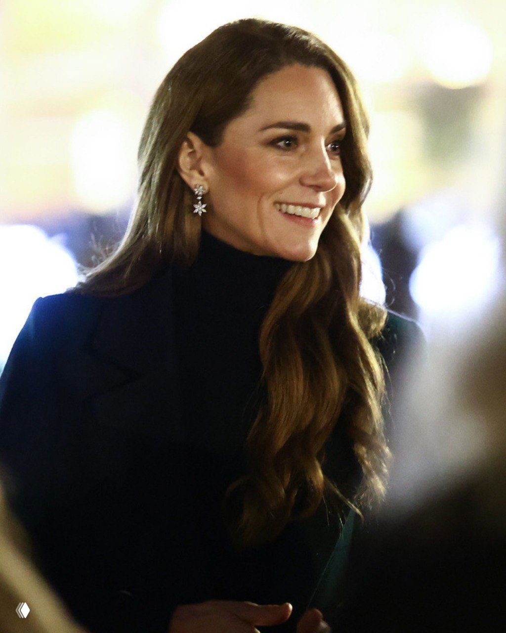 Крупный портрет принцессы Кейт в тёмном свитере и изящных серьгах, тёплая улыбка в вечерней подсветке благотворительного события.