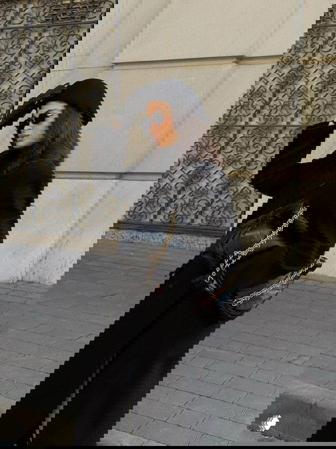 Женщина в тёмной одежде и шляпке-клош с солнцезащитными очками на городской улице — сочетание ретро и современного силуэта.