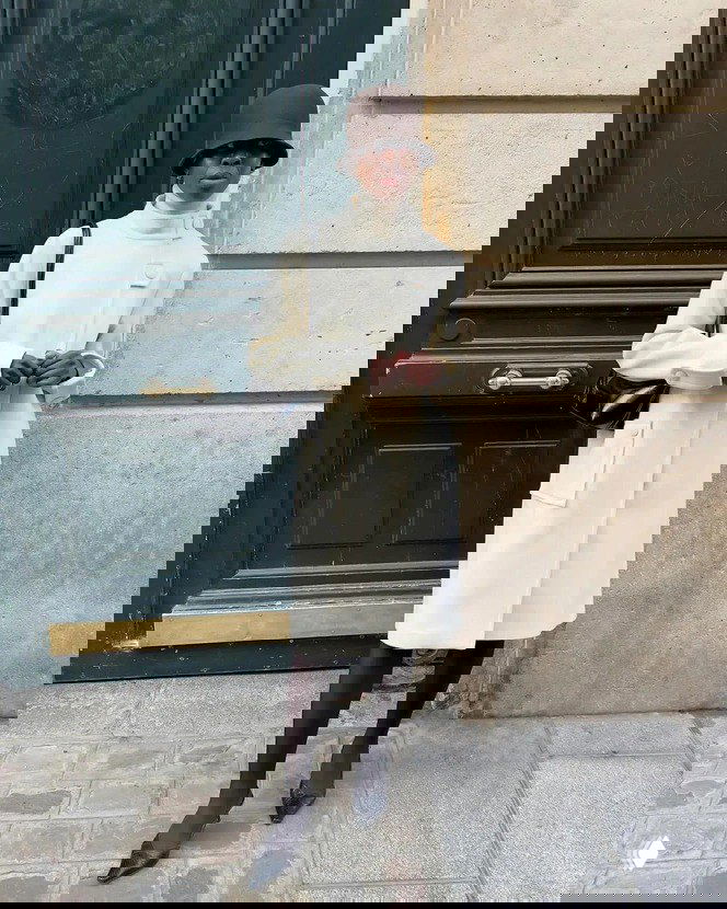 Модель в элегантном светлом пальто и тёмной шляпе-таблетке позирует у двери — сдержанный лаконичный образ с контрастом силуэтов.