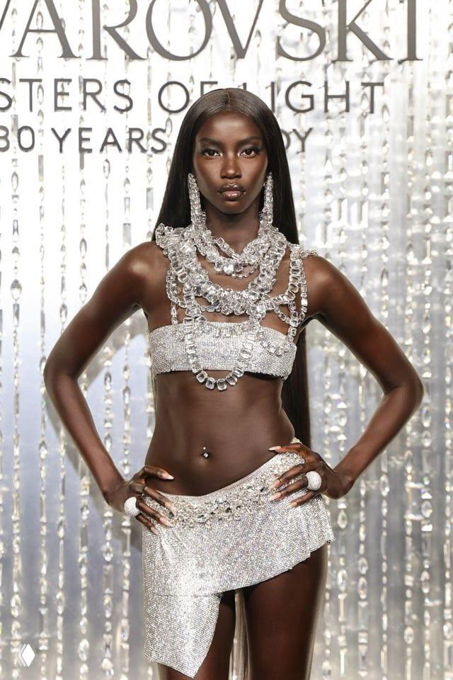 Темнокожая модель в серебристом мини‑ансамбле, усыпанном кристаллами Swarovski; поза в полный рост на фоне логотипа бренда.