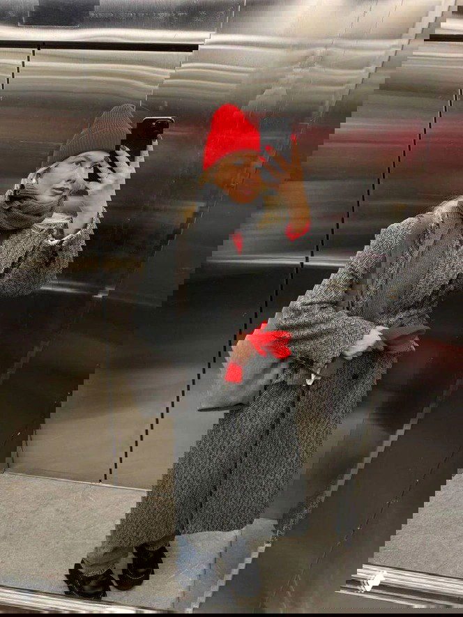 Селфи в зеркале: женщина в красной вязаной шапке и сером пальто показывает яркий повседневный вариант классической шапки с акцентным цветом.