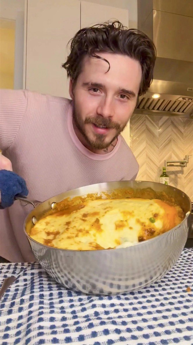 Мужчина на домашней кухне держит большую кастрюлю с запечённым блюдом; видна кухонная плитка, рабочая поверхность и посуда.