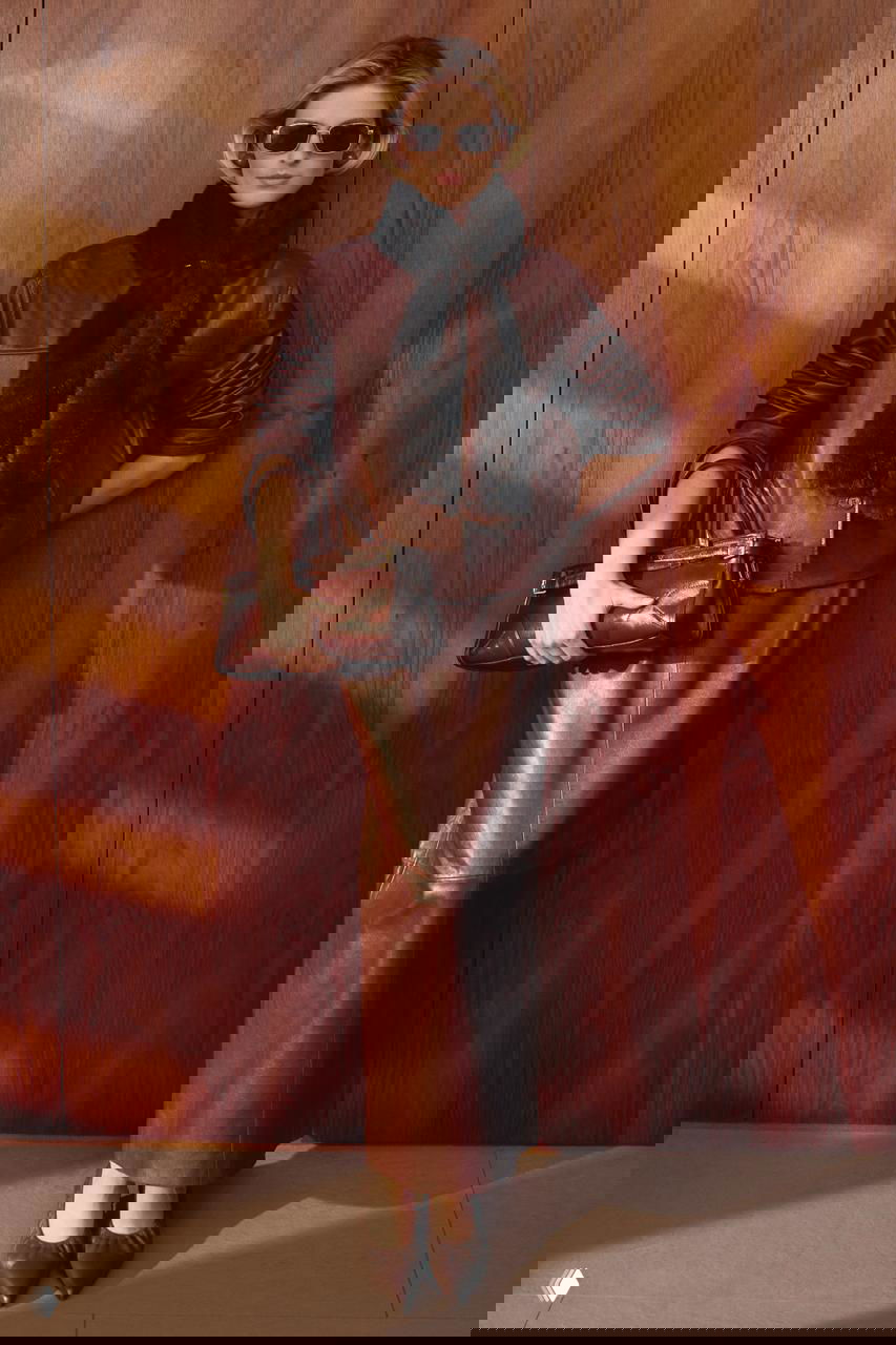 Модель в коричневом кожаном костюме: бомбер и длинная юбка, сумка в руках, солнцезащитные очки на фоне деревянной панели — фотосессия Marie Claire, акцент на текстуры и силуэты.