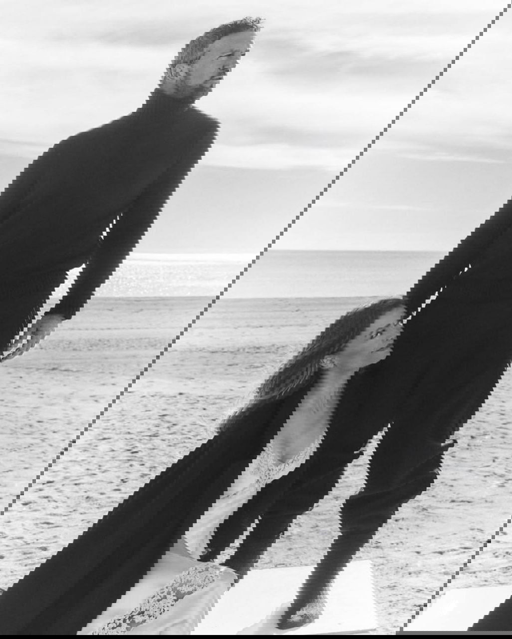 Николь Кимпел сидит у моря в платье, Антонио Бандерас стоит рядом на пляже — элегантный кадр из романтической фотосессии пары в минималистичных тонах.