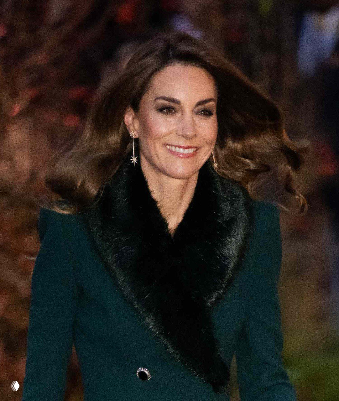 Женщина в изумрудном длинном пальто с тёмным меховым воротником улыбается на фоне вечерней подсветки у аббатства