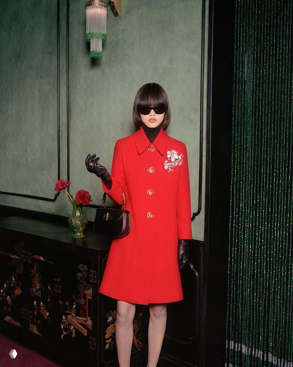 Женщина в красном пальто и тёмных очках стоит у зелёной стены рядом с вазой и декоративными элементами — образ из кампейна Gucci.