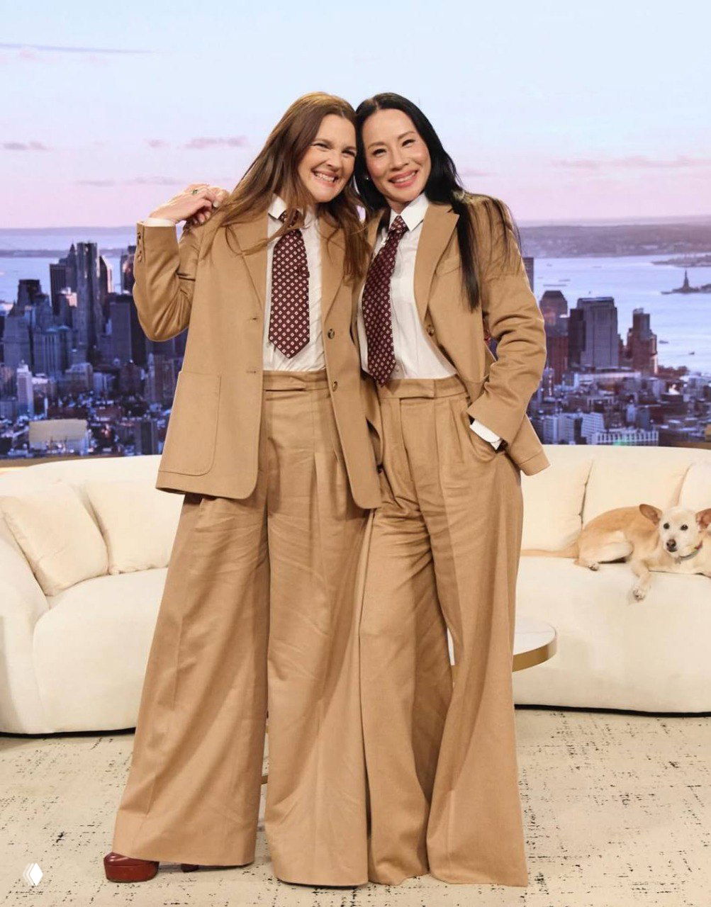 Две улыбающиеся женщины в бежевых костюмах позируют в студии на диване на фоне городского пейзажа; рядом видна собака