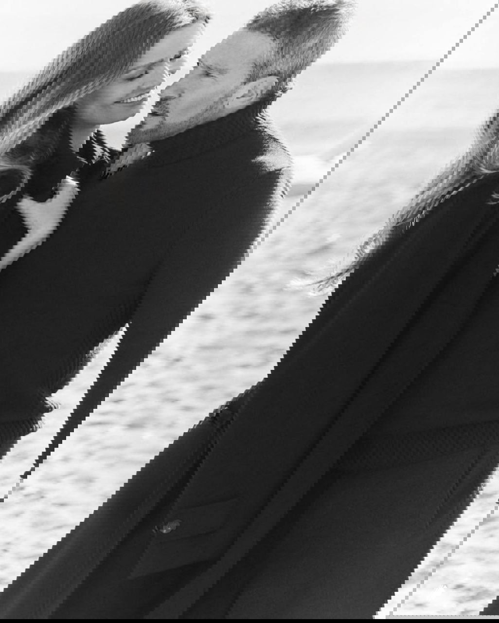 Крупный чёрно‑белый портрет: Николь Кимпел и Антонио Бандерас в пальто и свитере, обмениваются нежными взглядами на побережье.