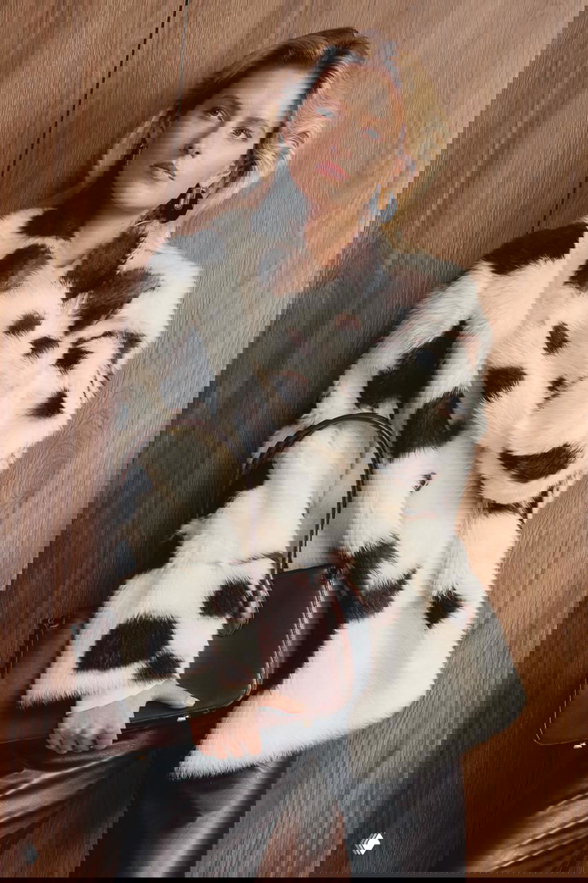 Модель в белой шубе с тёмно-коричневыми пятнами, тёмные кожаные брюки и лаконичная сумка на фоне деревянной панели — фотосессия Marie Claire, заметен акцент на анималистичные принты и контраст текстур.