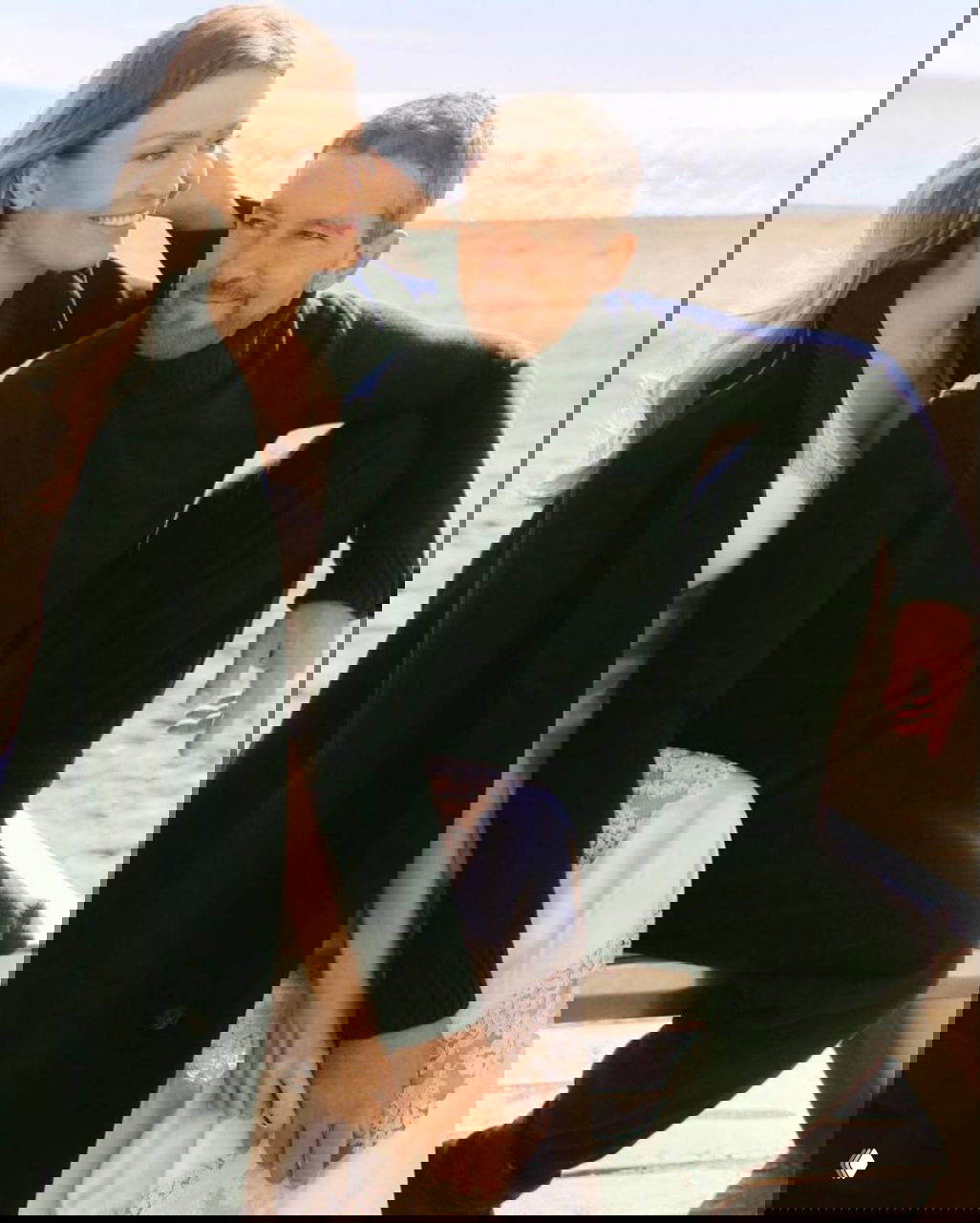 Цветной кадр фотосессии: Николь Кимпел и Антонио Бандерас сидят на скамье у моря, улыбаются и выглядят расслабленными в повседневных образах.