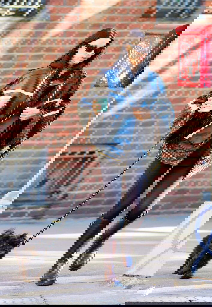 Женщина в чёрной кожаной куртке идёт по тротуару Нью-Йорка, держит стакан кофе и маленькую тёмную собаку на поводке.