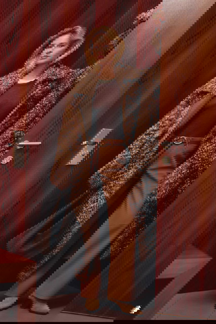 Модель в объёмной коричневой шубе из кудрявой шерсти, широкие брюки и большая замшевая сумка у дверного проёма — фотосессия Marie Claire, игра с пропорциями и фактурами в фокусе.