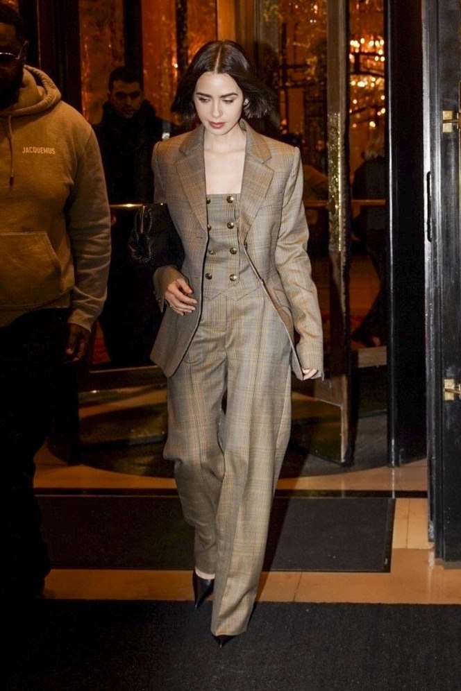 Лили Коллинз в выдержанном клетчатом двубортном костюме и широких брюках выходит из здания в вечернем освещении — элегантный деловой образ.