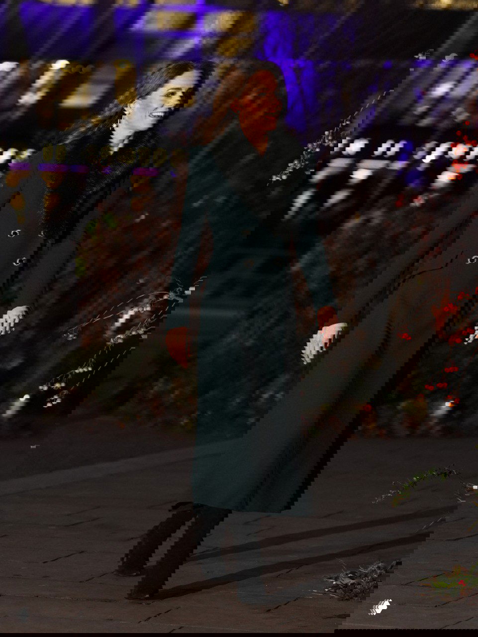 Женщина в длинном тёмно-зелёном пальто идёт по дорожке в вечерней подсветке перед зданием с праздничными огнями