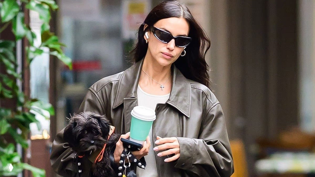 Крупный план женщины в очках с маленькой собакой на руках и бумажным стаканом на фоне улицы Нью-Йорка.