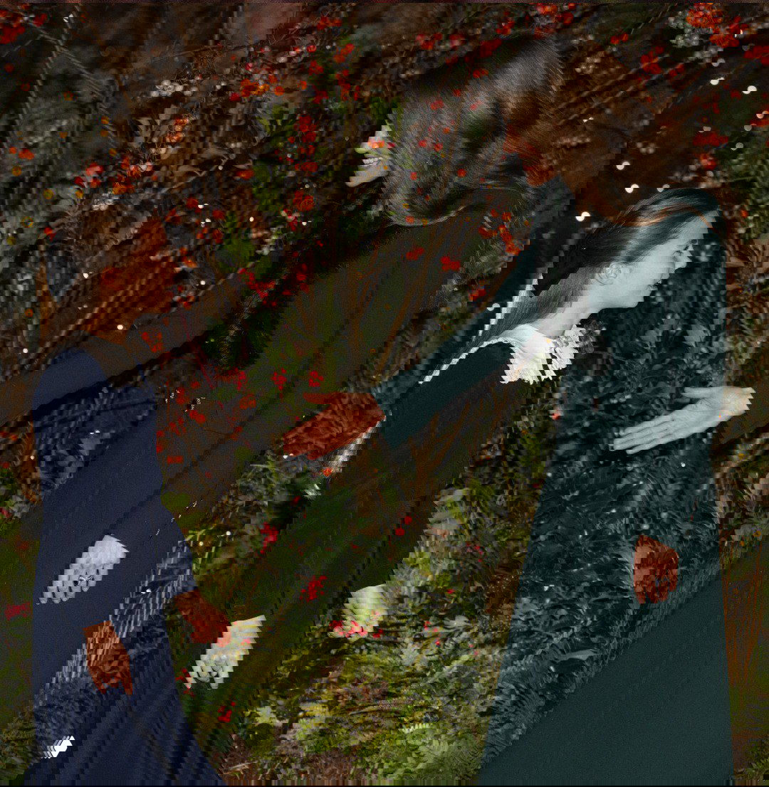 Женщина в изумрудном пальто протягивает руку и здоровается с девочкой на фоне праздничного декора и огней