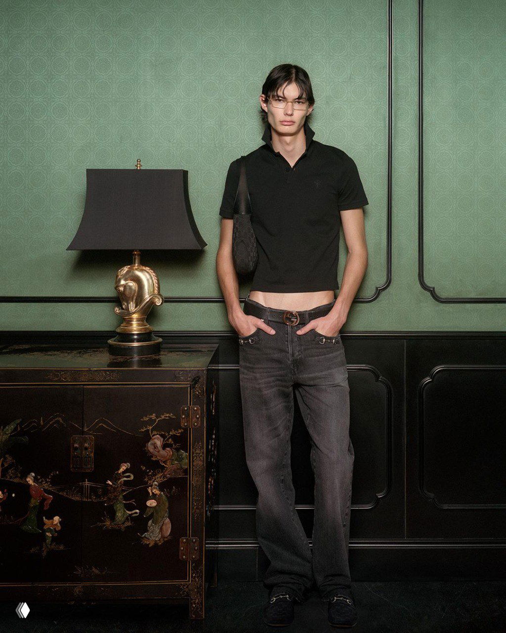 Молодой мужчина в тёмной футболке и джинсах стоит у комода с восточным орнаментом на фоне зелёной стены — рекламный образ Gucci The Gathering.