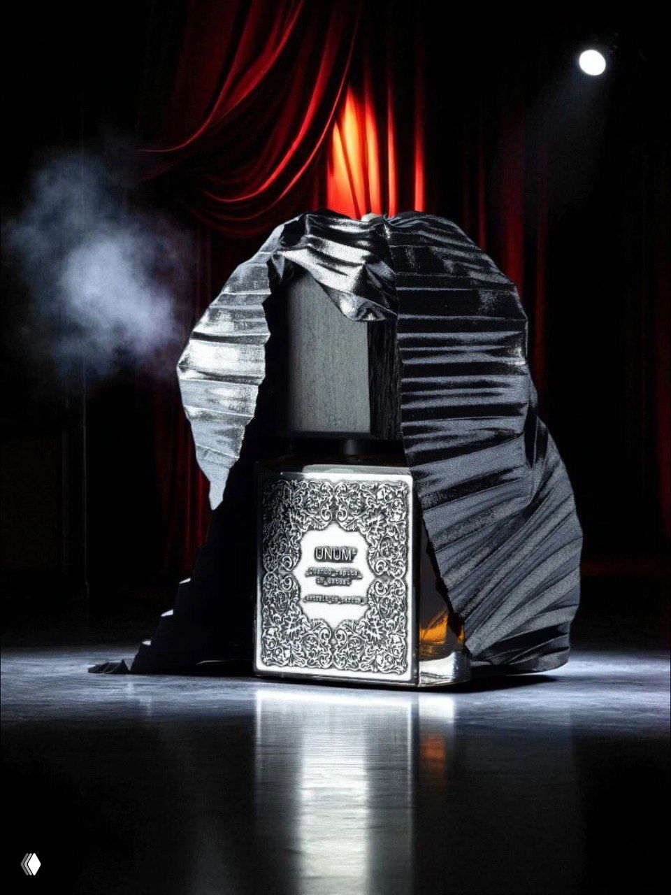 Флакон парфюма в театральной постановке: металлическая, орнаментированная коробка и тёмная ткань на сцене с прожекторным светом и дымкой.
