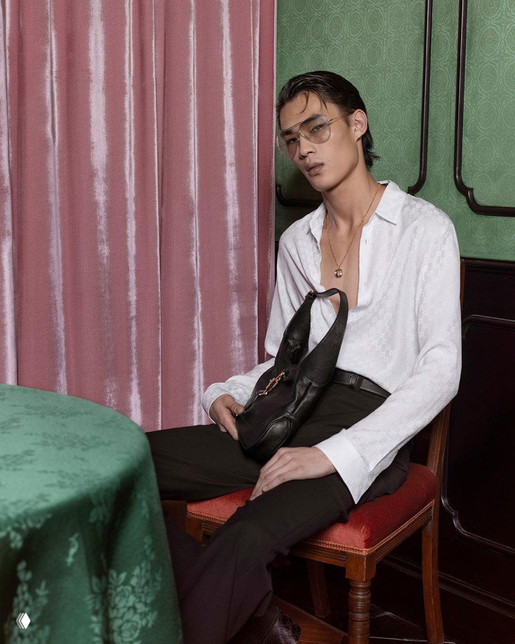 Модель сидит у стола в белой открытой рубашке, в руках сумка Gucci; кадр подчёркивает аксессуар и атмосферу кампейна The Gathering.
