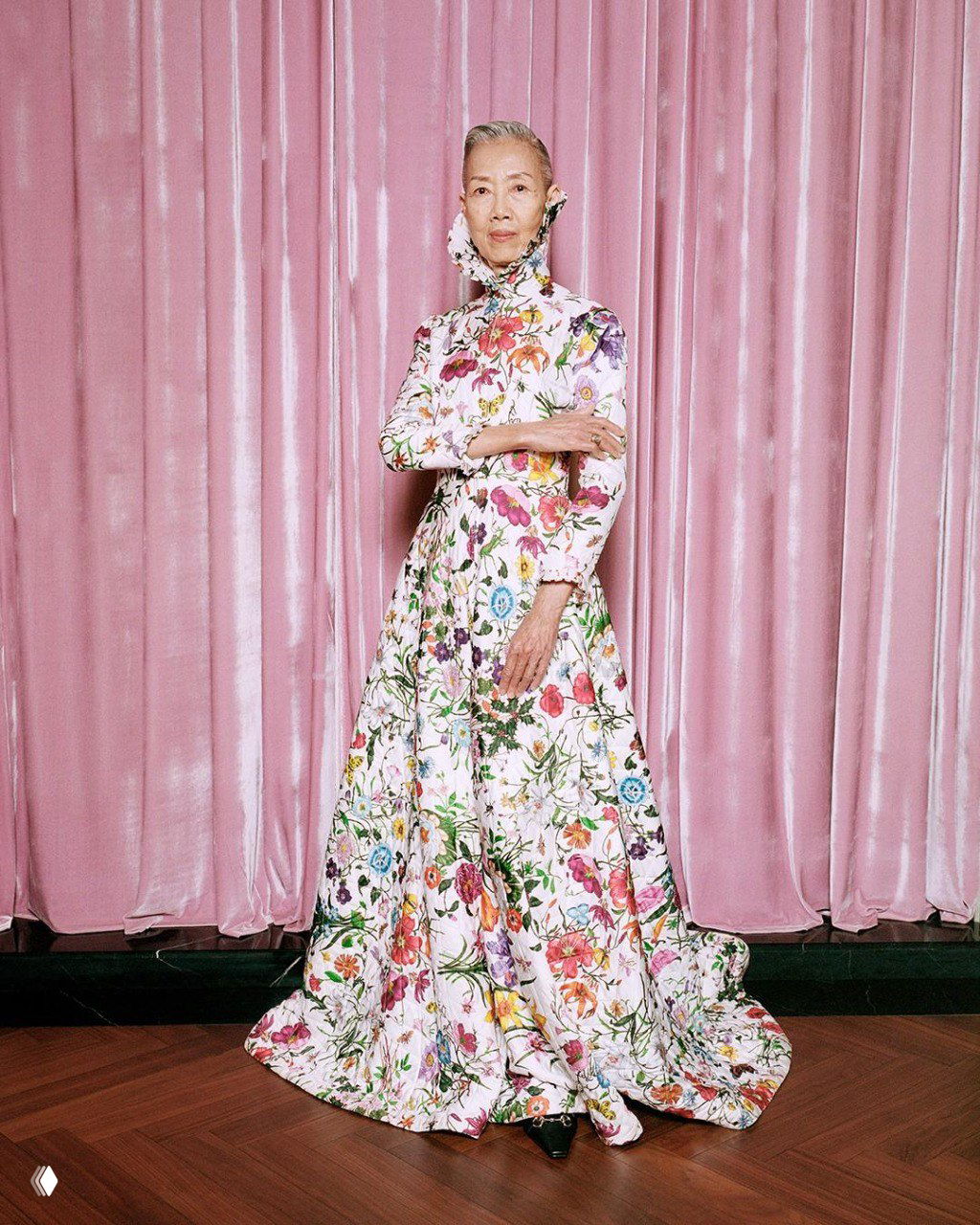 Пожилая женщина в длинном цветочном платье позирует на фоне розовых штор — кадр, подчёркивающий многообразие поколений в кампейне Gucci The Gathering.