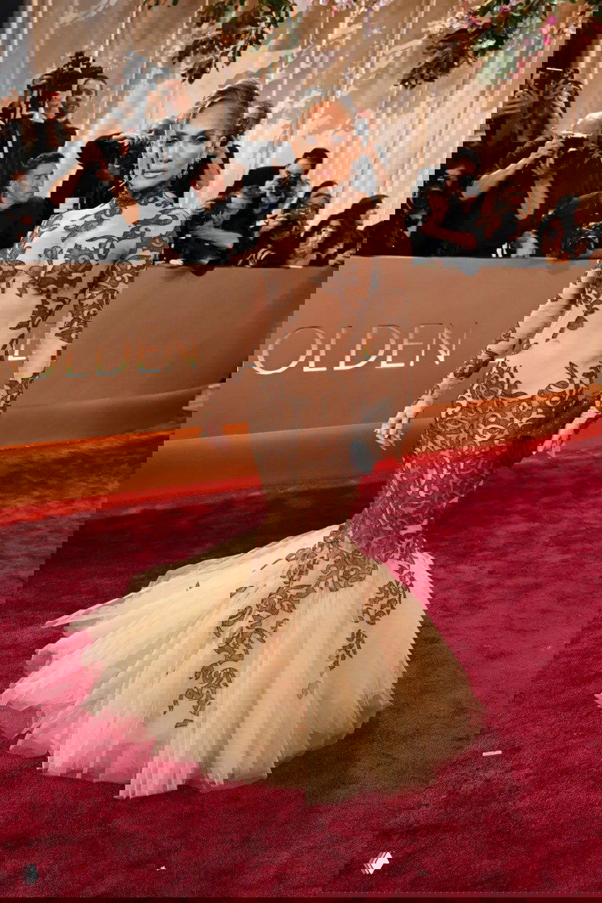 Наряды нюдовой гаммы: облегающее платье с прозрачными вставками и многослойной юбкой‑русалкой на красной дорожке на фоне логотипа мероприятия.