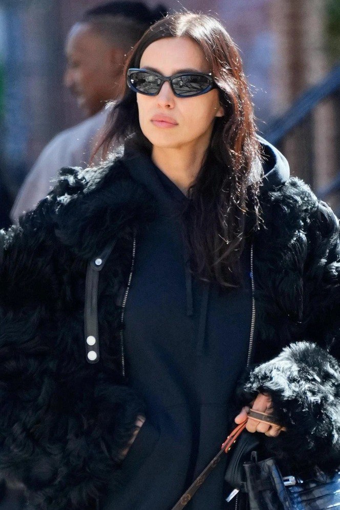 Крупный план: объёмная чёрная меховая или фактурная куртка, худи и узкие солнцезащитные очки в ретро‑стиле на городском фоне.