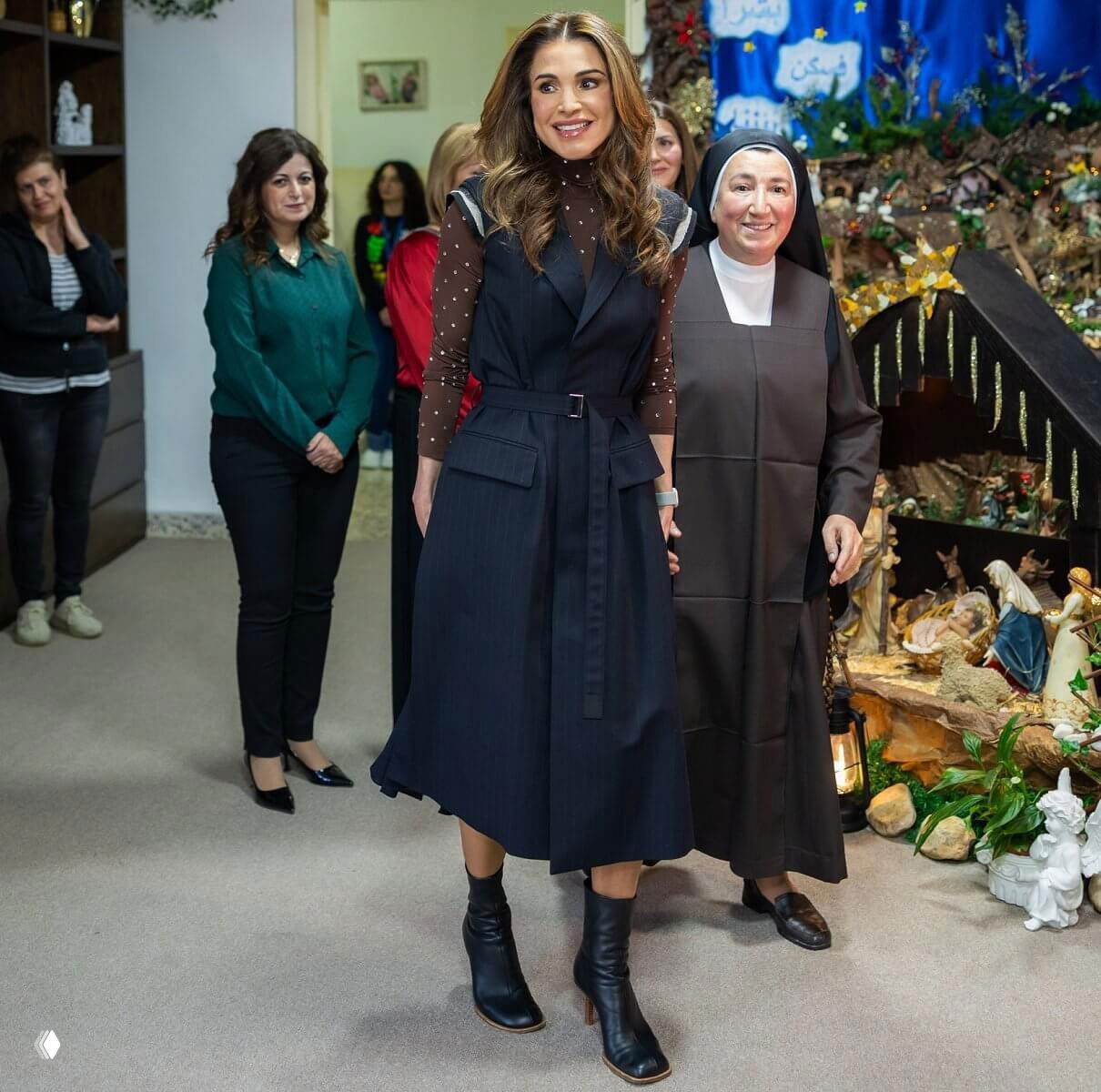 Королева Рания идёт по залу центра рядом с монахиней; на заднем плане рождественские декорации и люди, наблюдающие за визитом.