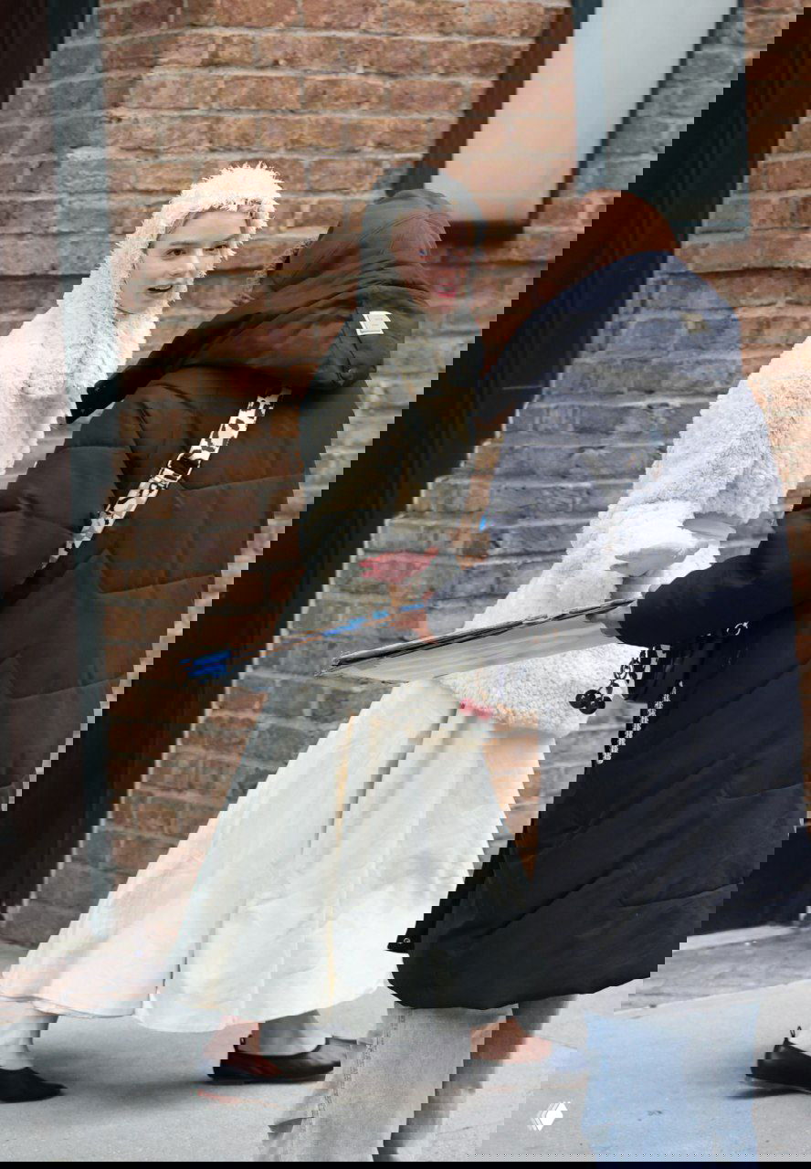 Папарацци: женщина в кремовой плюшевой шубе и длинной юбке идёт по городу у кирпичной стены, уличный зимний кадр.