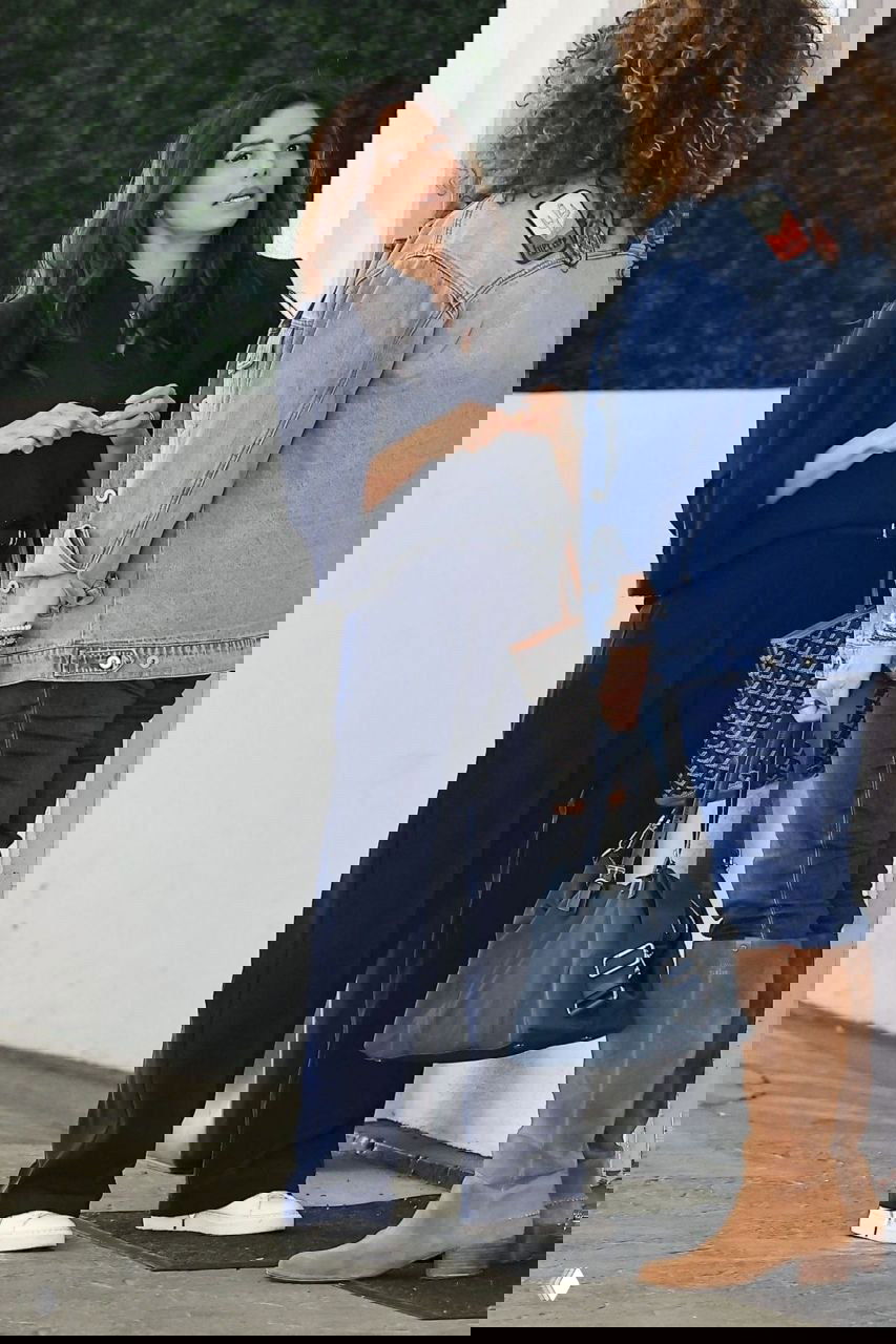 Две женщины разговаривают у входа: одна в тёмно-синем костюме и белых кроссовках, вторая в джинсовой куртке и сапогах; городской фон.