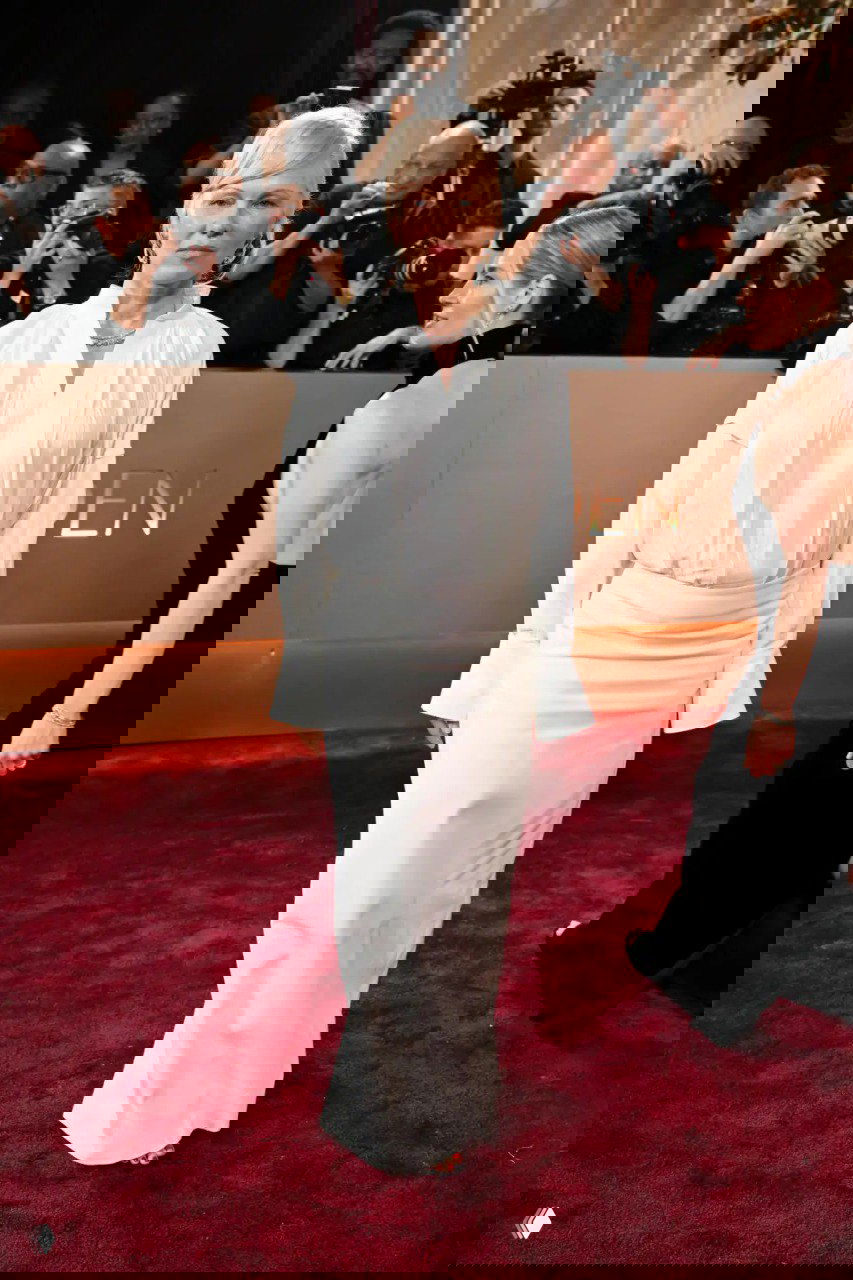 Женщина в строгом белом костюме и юбке стоит на красном ковре, вокруг нее — фотографы и другие гости церемонии, спокойный элегантный образ.