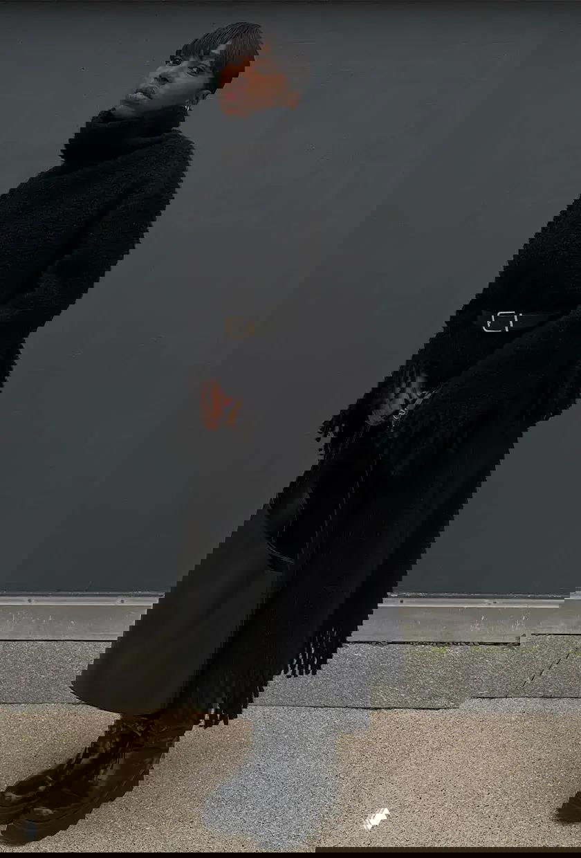 Аутфит в тёмных тонах: объёмная чёрная водолазка с поясом, длинное пальто и грубые ботинки на ровном фоне городской стены.