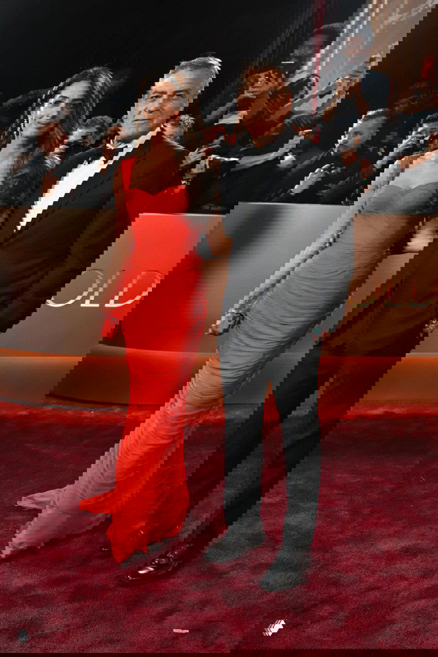 Пара в вечерних нарядах — женщина в ярком красном платье и мужчина в классическом смокинге на фоне декора церемонии и фотографов.