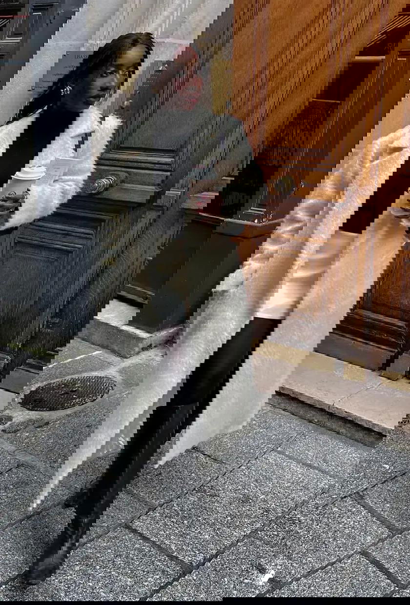 Образ в светлом пальто и чёрной водолазке: мини-юбка, высокие сапоги и кофе в руке на фоне деревянной двери.