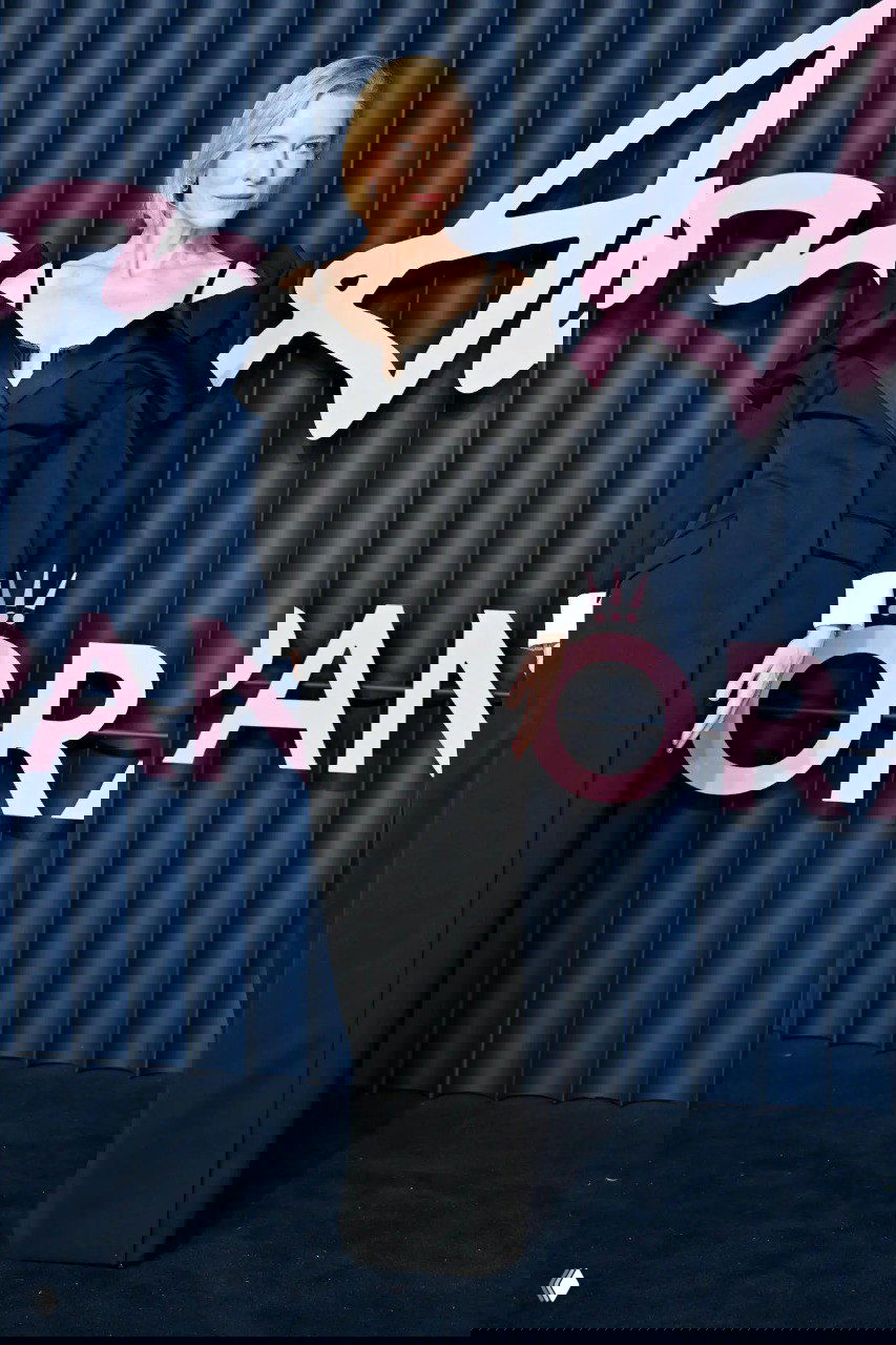 Портрет знаменитости в длинном чёрном вечернем платье с открытыми плечами на фоне синей стенки и крупного розового логотипа мероприятия.