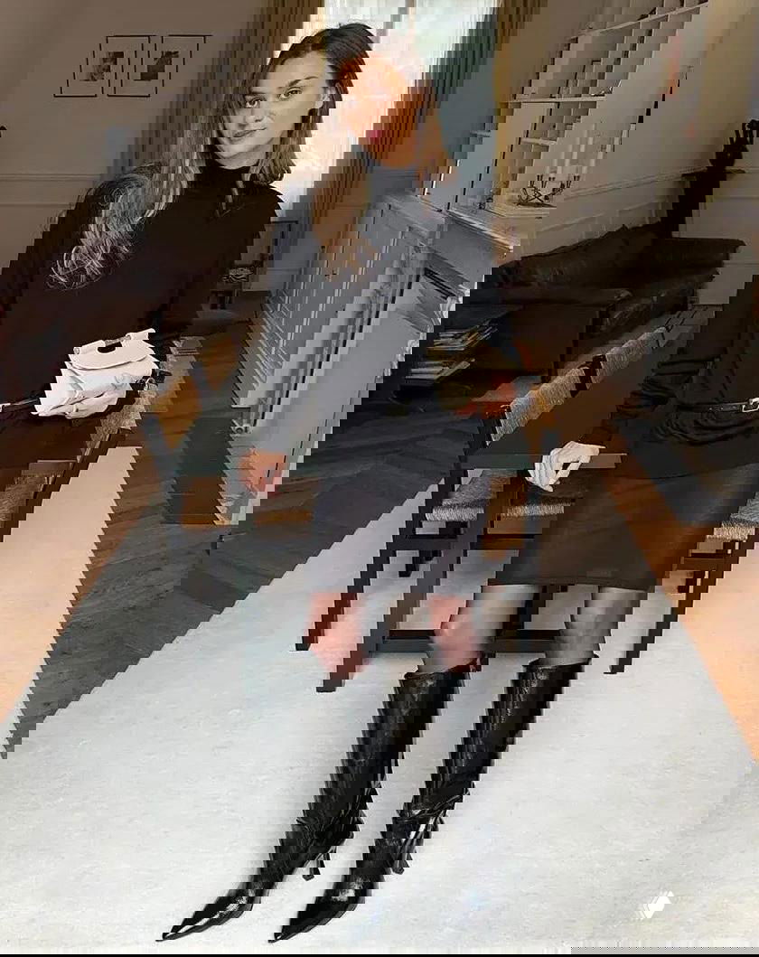 Женщина в чёрной водолазке, кожаной юбке-миди и высоких лакированных сапогах, с кремовым клатчем на фоне столовой и стола.