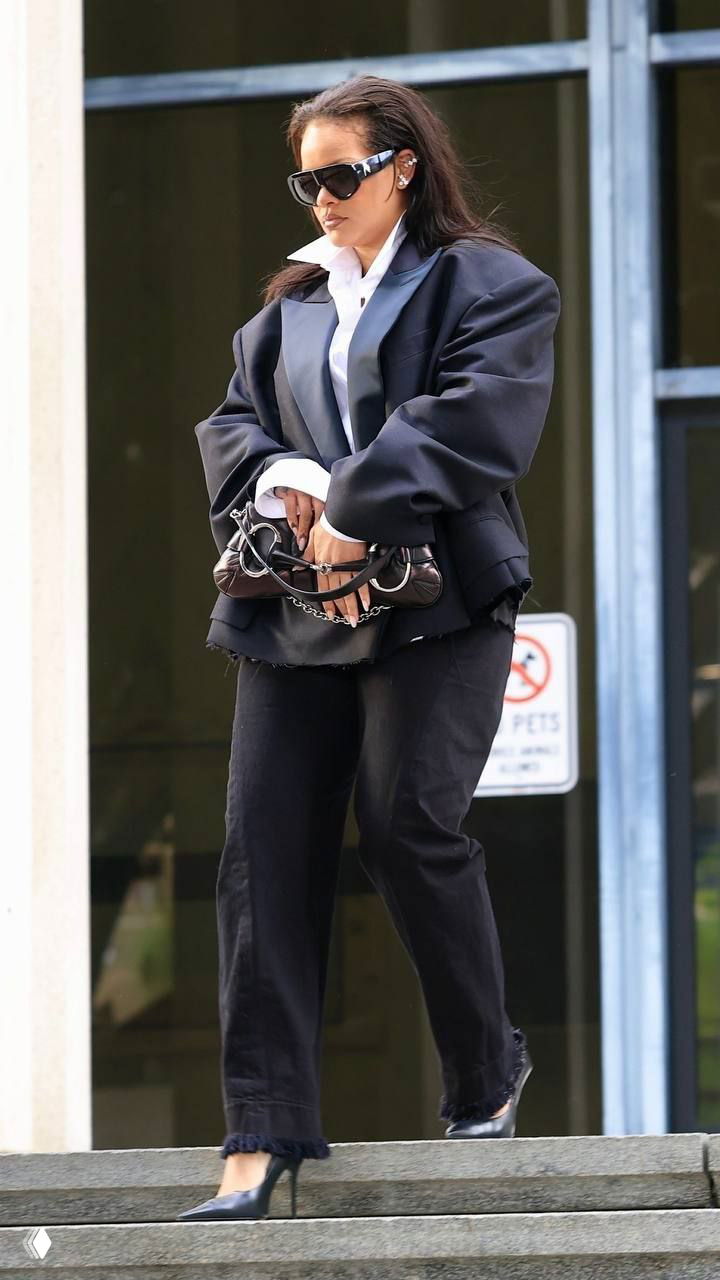 Днёвной уличный кадр: женщина в свободном тёмном пиджаке и солнцезащитных очках спускается по ступеням у входа в здание.