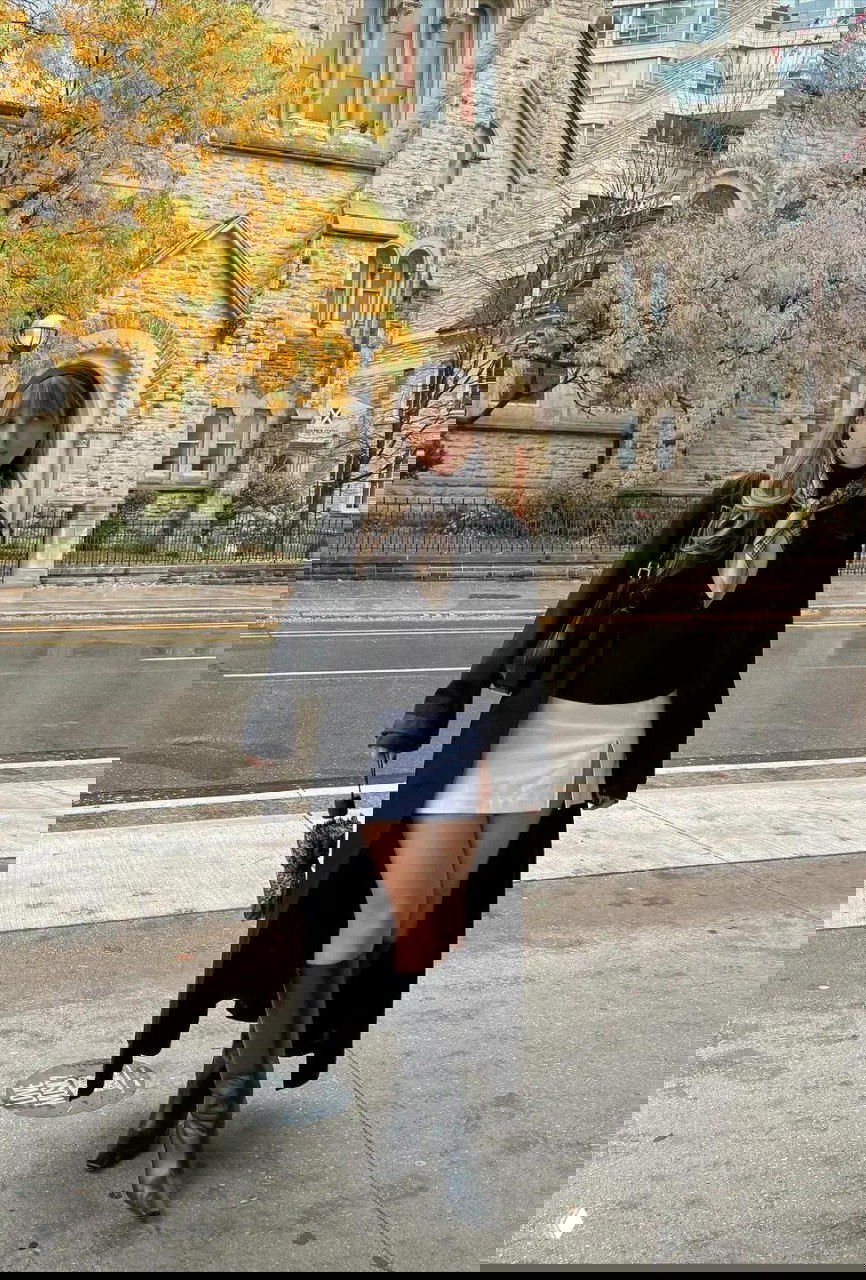 Уличный снимок: чёрная водолазка в сочетании с длинным чёрным пальто и белой юбкой-мини, высокие сапоги и осенний городской фон.