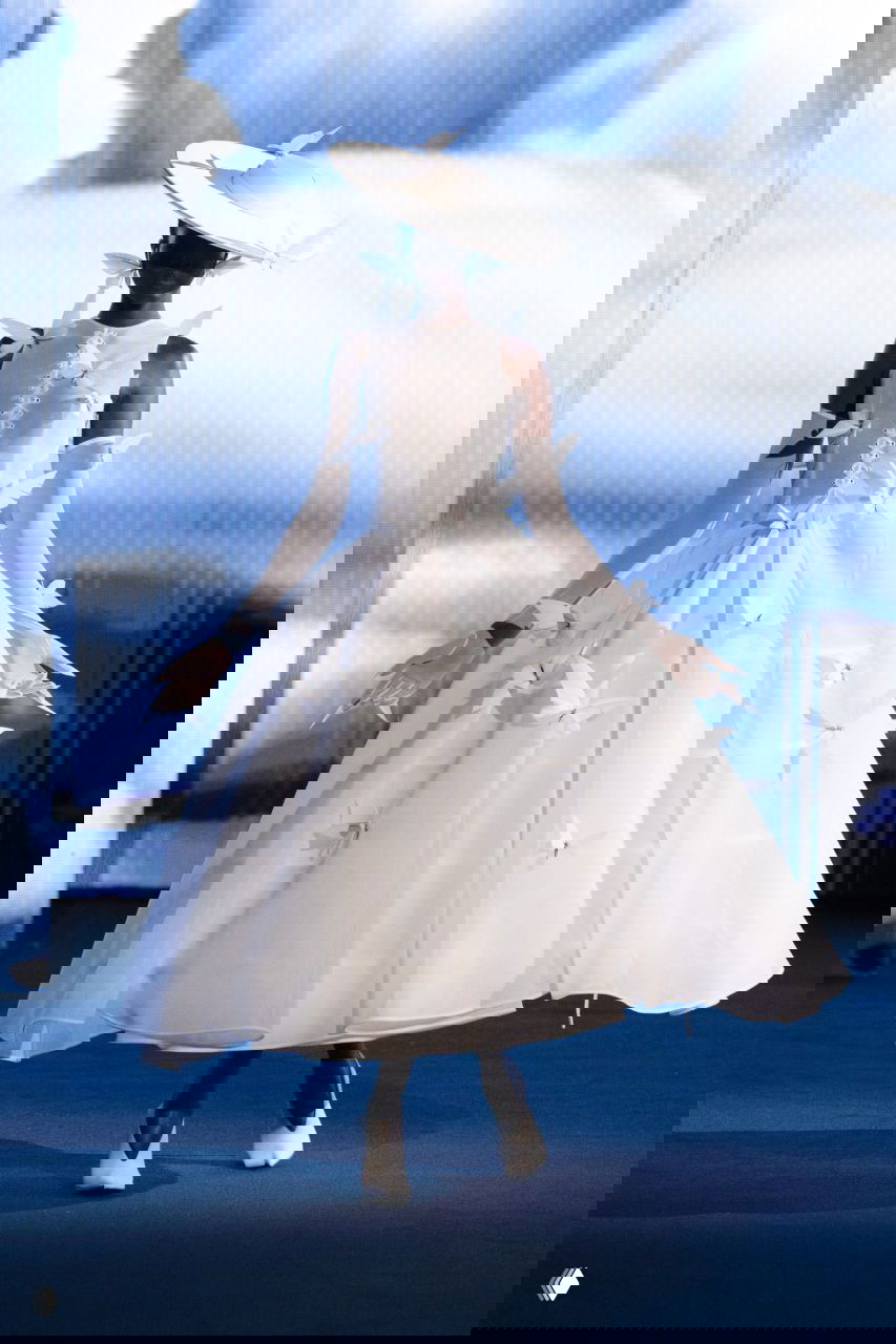 Белое платье-кринолин Роберта Вуна с объёмной юбкой и декором в виде бумажных мотыльков, модель в шляпе на фоне облачного видеоэкрана.