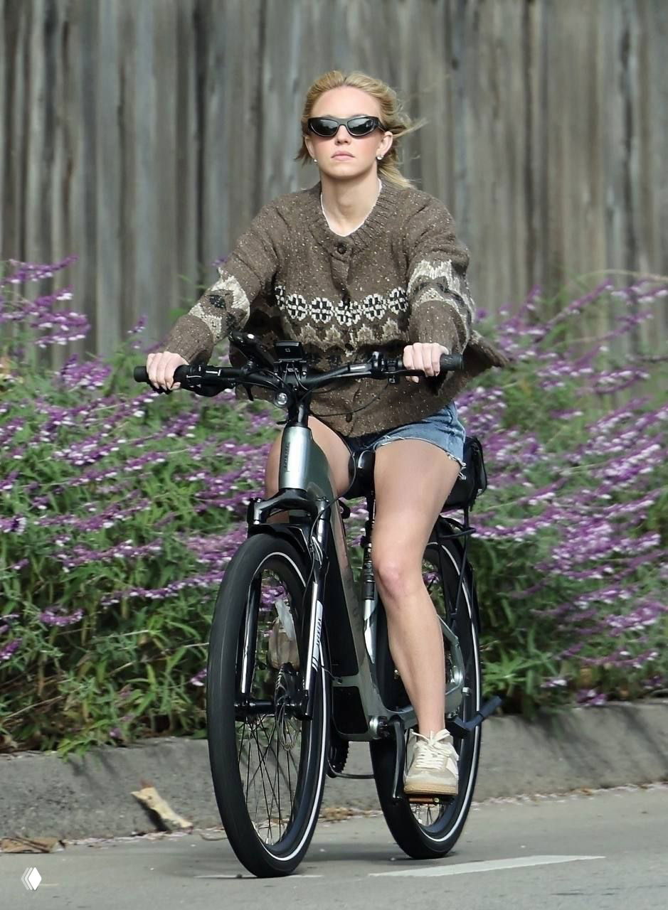 Папарацци: женщина на велосипеде в коричневом вязаном кардигане и джинсовых шортах едет по улице, вдоль бордюра фиолетовые цветы.