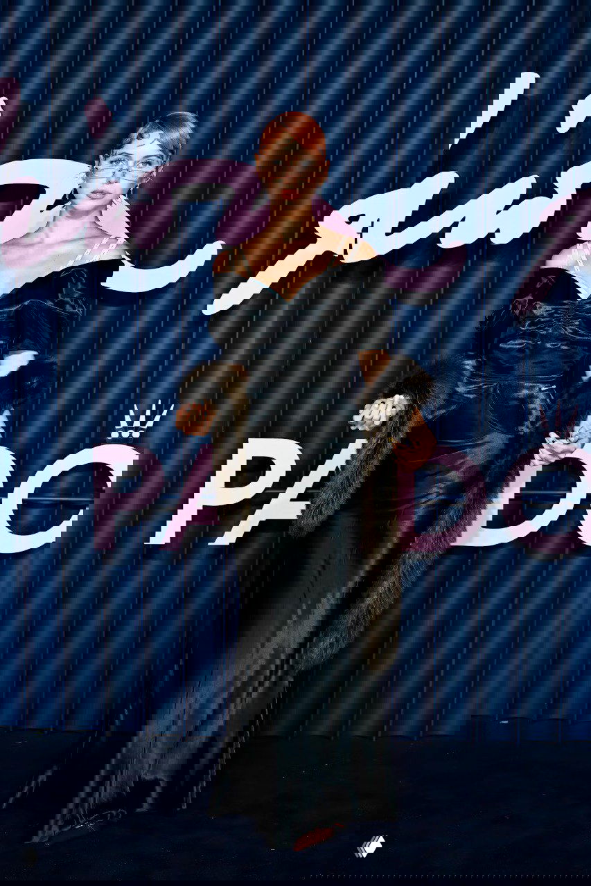 Фигура в чёрном атласном платье с меховой накидкой и тонкими бретелями на фоне синего декора с розовой типографикой бренда.