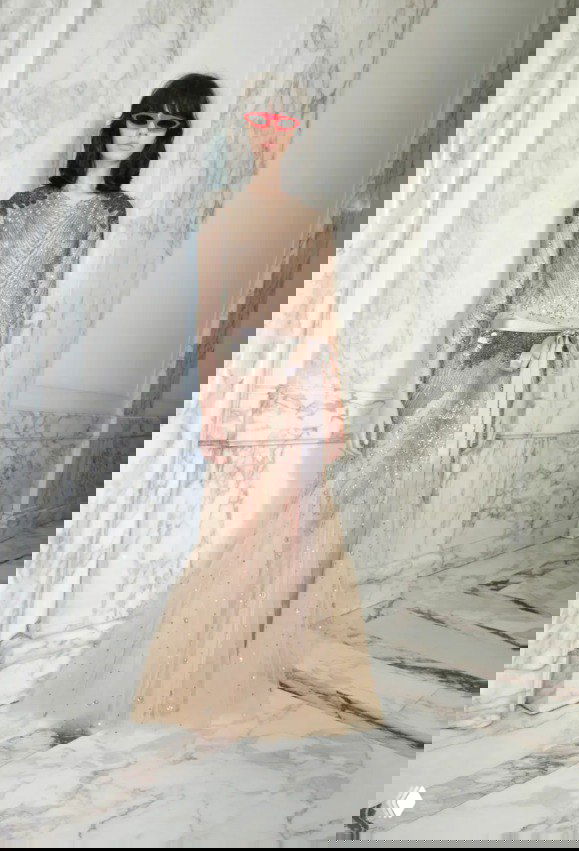 Стройная модель в прозрачном длинном платье с блёстками и тонкой поясной лентой на мраморном фоне — деталь кутюрной эстетики Valentino.