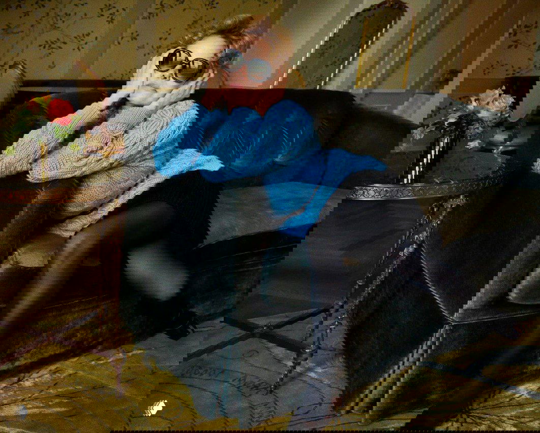 Интерьерный снимок женщины в голубом вязаном свитере и солнцезащитных очках, расслабленная поза на диване в уютной комнате с цветочным акцентом на столике.