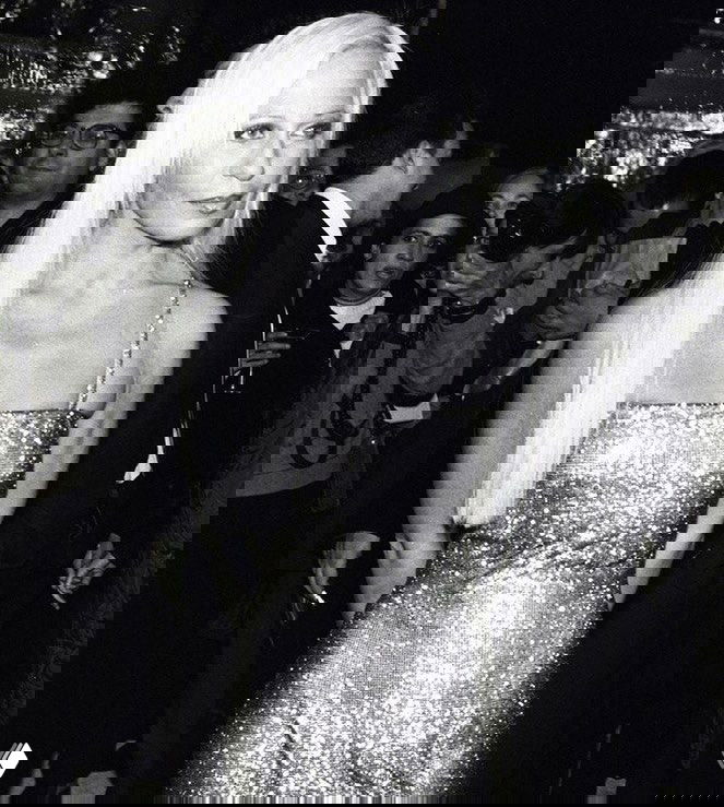 Чёрно‑белое фото: женщина в блестящем вечернем платье на светском мероприятии, окружённая фотографами и гостями, портретный ракурс.