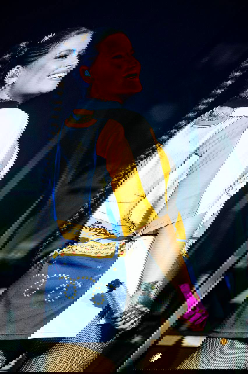 Нелли Фуртадо в профиль на сцене: улыбается, держит блестящий микрофон, длинная коса и заметный сценический образ с яркими цветами и аксессуарами.