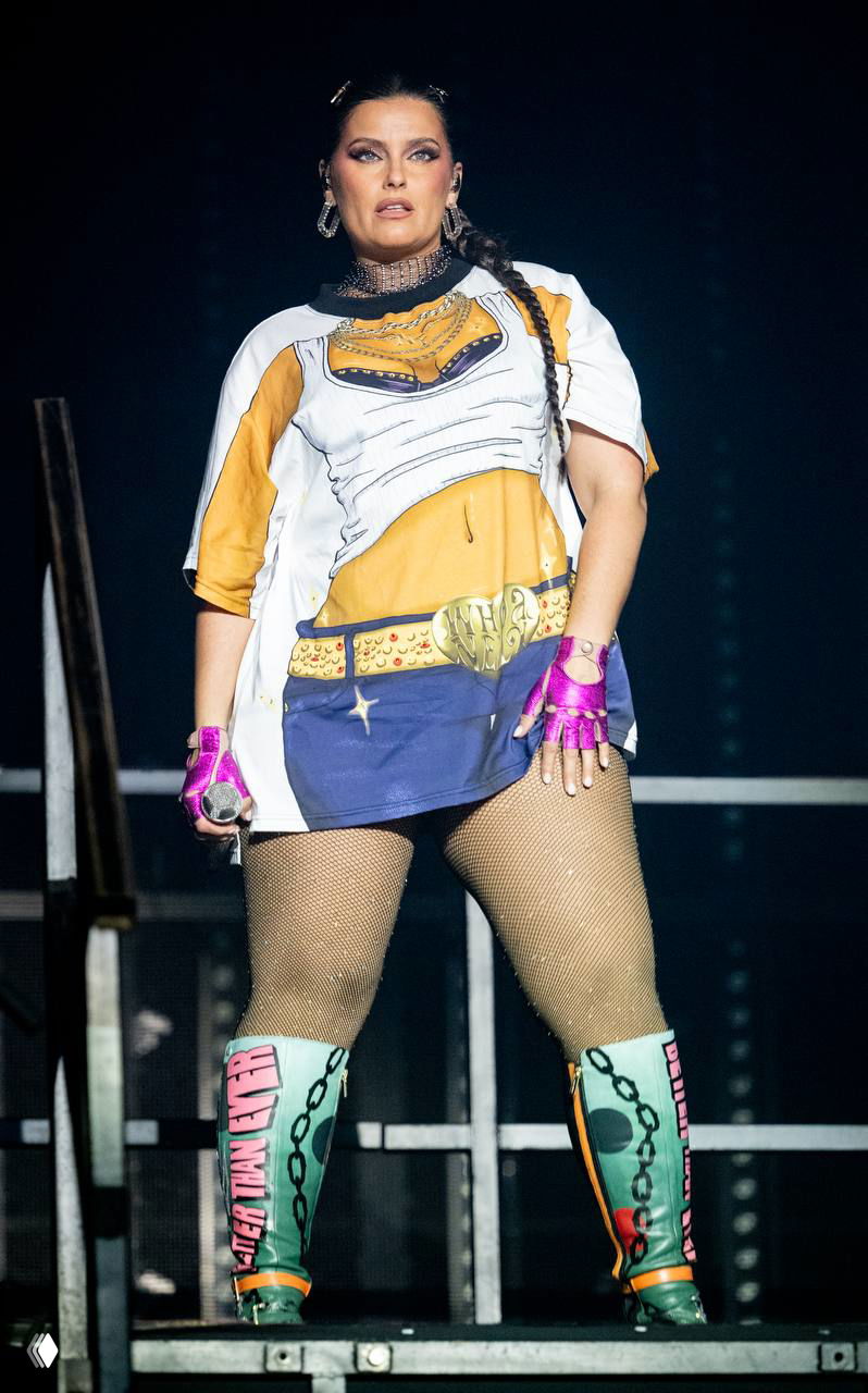 Крупный план Нелли Фуртадо в сценическом костюме и сеточных колготках: серьёзное лицо, яркие аксессуары и концертный свет вокруг неё.