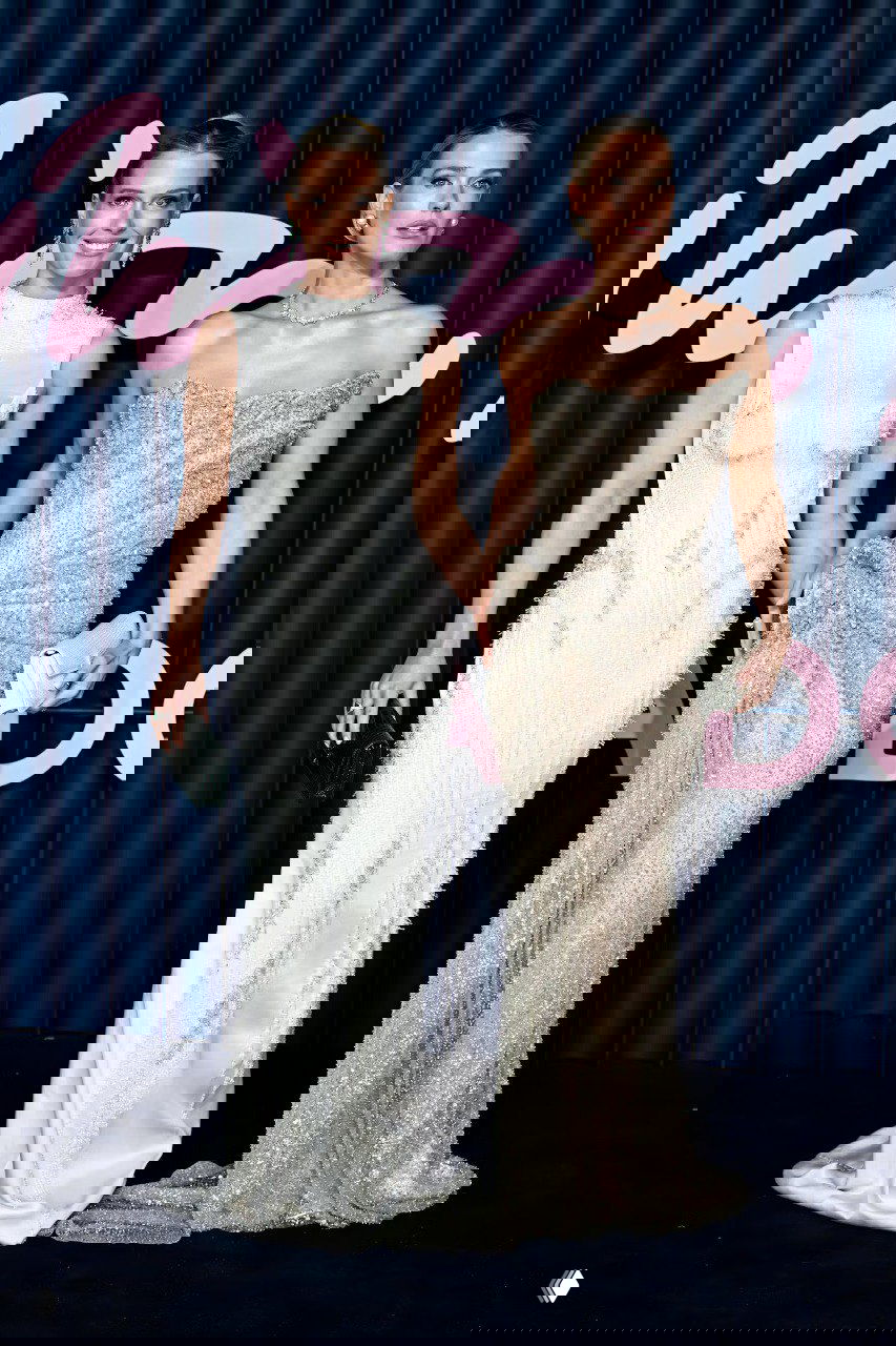 Две женщины в длинных вечерних платьях — одно белое, другое блестящее — стоят вместе на фоне синего баннера с розовой надписью мероприятия.