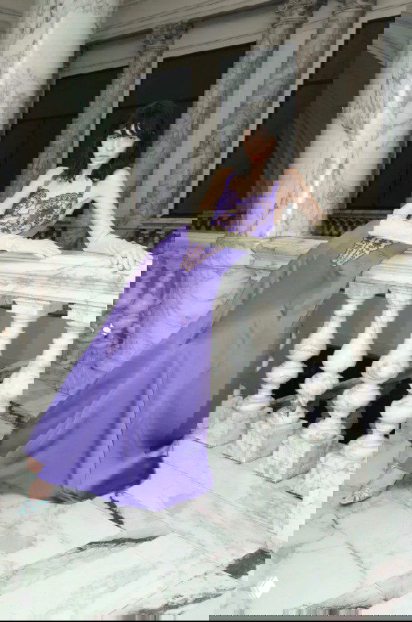 Модель в сиреневом атласном платье с кружевной вставкой опирается на мраморный парапет — элегантный вечерний образ из Pre‑Fall 2026.