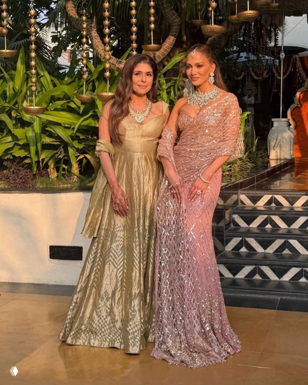Дженнифер Лопес в нежно-розовом сари рядом с другой женщиной на фоне зелёной тропической декорации и подвесных светильников свадьбы в Индии.