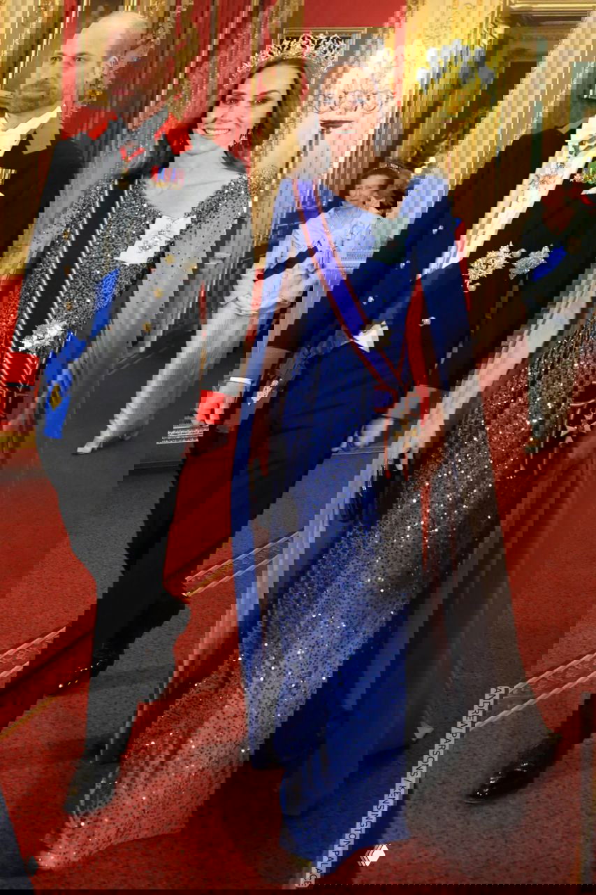 Женщина в длинном синем платье с накидкой и тиарой идёт рядом с мужчиной в парадном костюме по коридору Виндзорского замка.