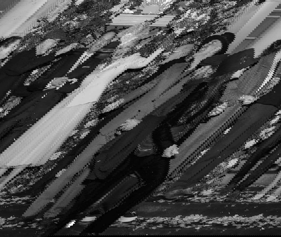 Широкий черно‑белый кадр: пара выходит под арку из цветов, гости вокруг бросают лепестки — праздничная уличная сцена после церемонии.